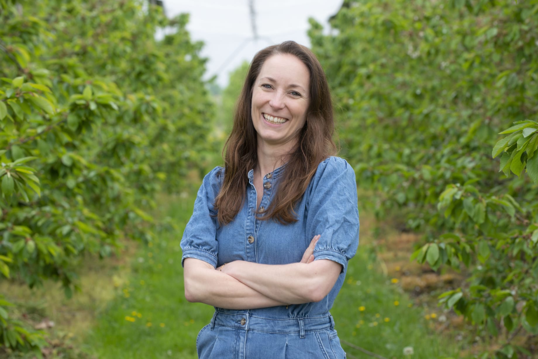 Nicole staat met haar armen over elkaar, lacht, draagt een spijkerstof pak en staat in een veld met bomen