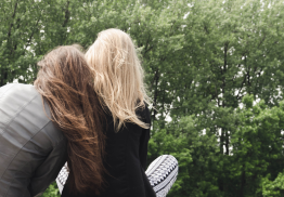 Twee vrouwen zitten naast elkaar, rug naar het beeld. Vrouw met bruin lang haar heeft hoofd op schouder van vrouw met blond halflang haar. Ze zitten in het gras op de grond. je ziet bomen op het achtergrond.