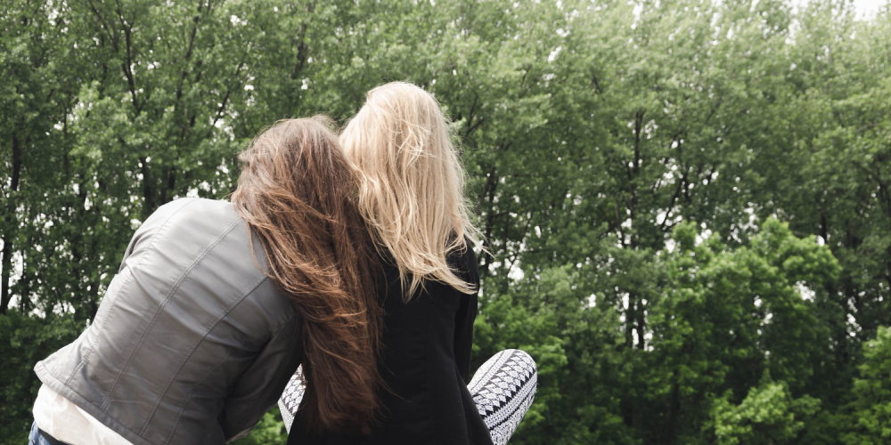 Twee vrouwen zitten naast elkaar, rug naar het beeld. Vrouw met bruin lang haar heeft hoofd op schouder van vrouw met blond halflang haar. Ze zitten in het gras op de grond. je ziet bomen op het achtergrond.